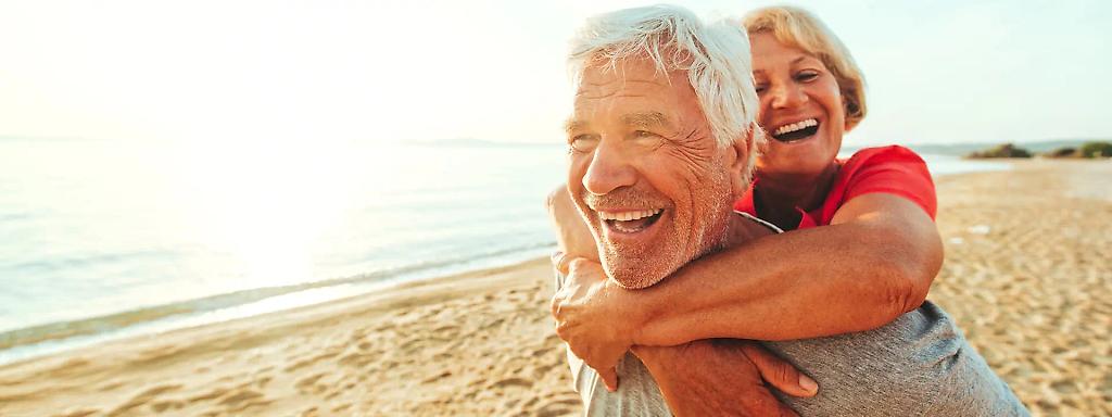 Älters Paar am Strand, Frau umarmt Mann von hinten. Beide lachen glücklich